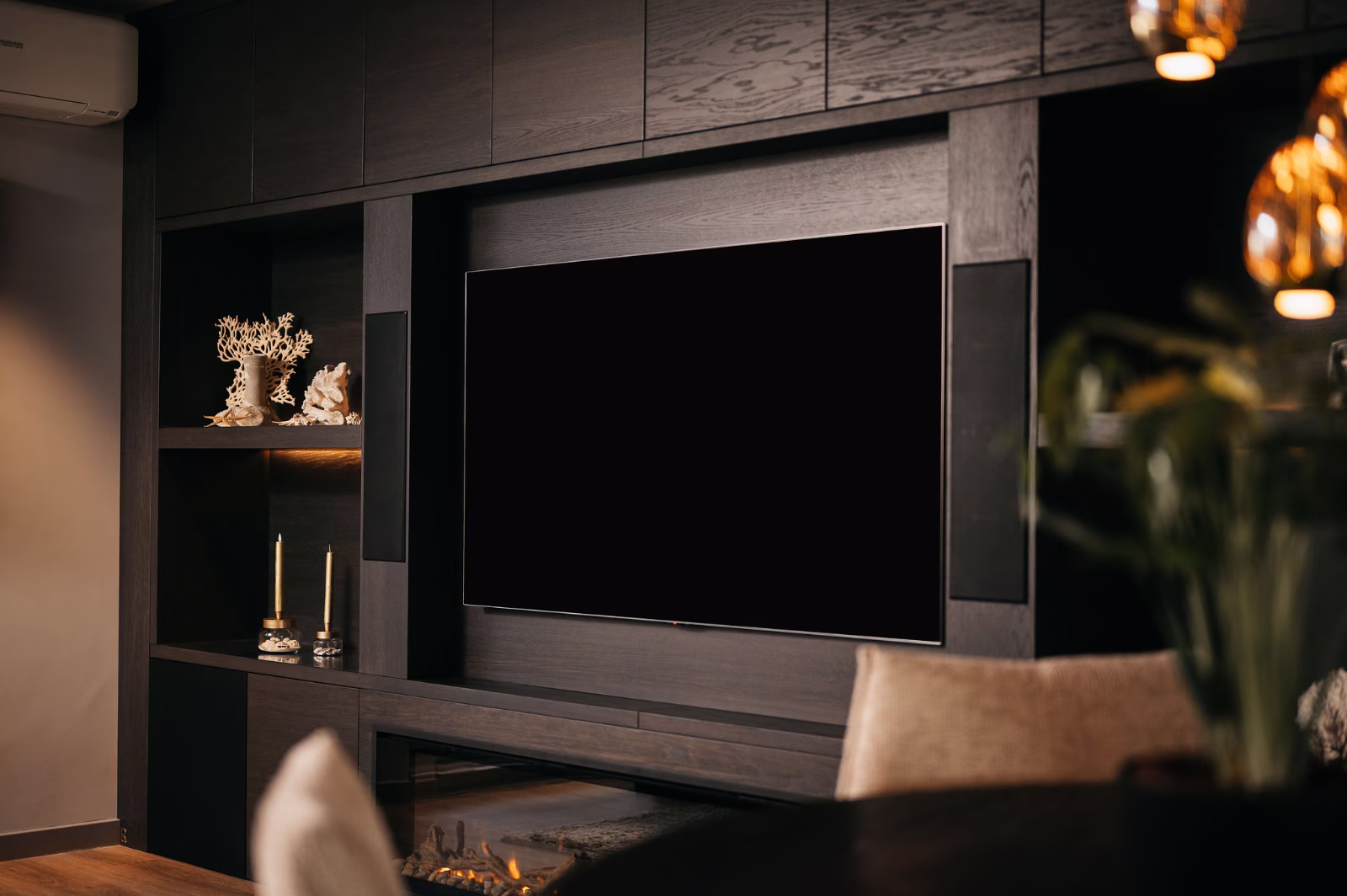 Een moderne woonkamer met een grote flatscreentelevisie op een donkerhouten wandmeubel, decoratieve voorwerpen en kaarsen op planken en een vage eettafel en stoel op de voorgrond.