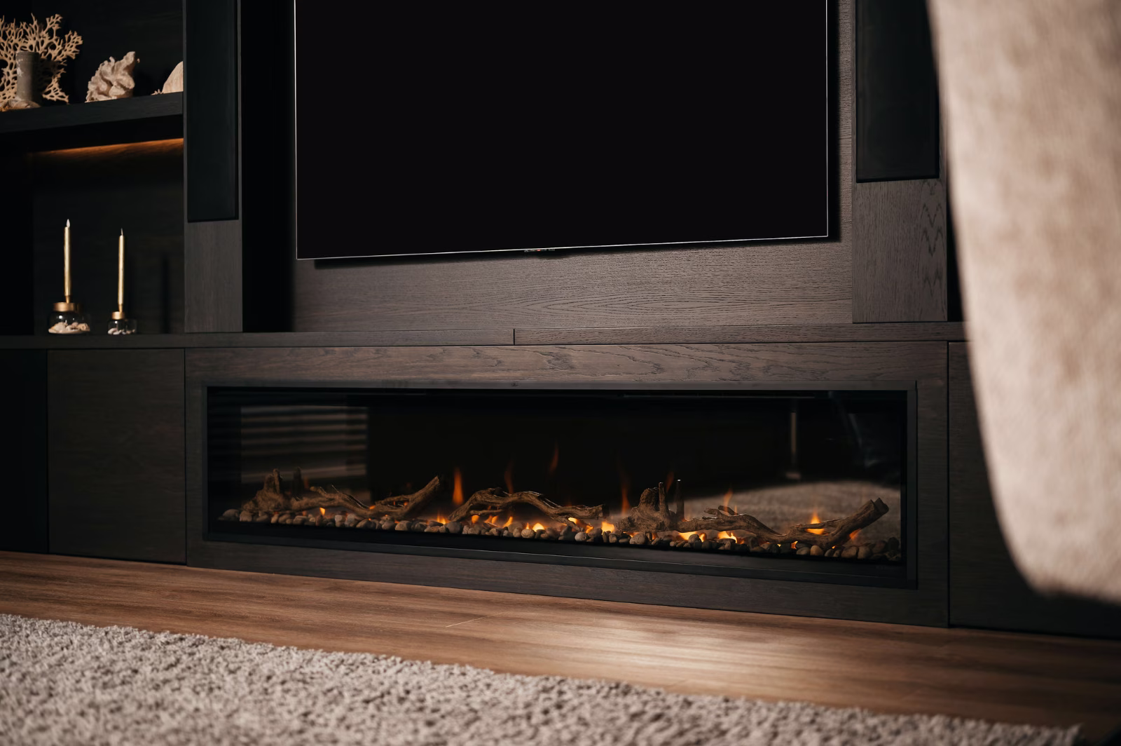 Een moderne elektrische open haard met een glazen voorkant, met gloeiende sintels en kunstmatige houtblokken, is ingebouwd in een donkerhouten entertainmentmeubel onder een grote flatscreentelevisie. Kaarsen en decor zijn zichtbaar op nabijgelegen planken.