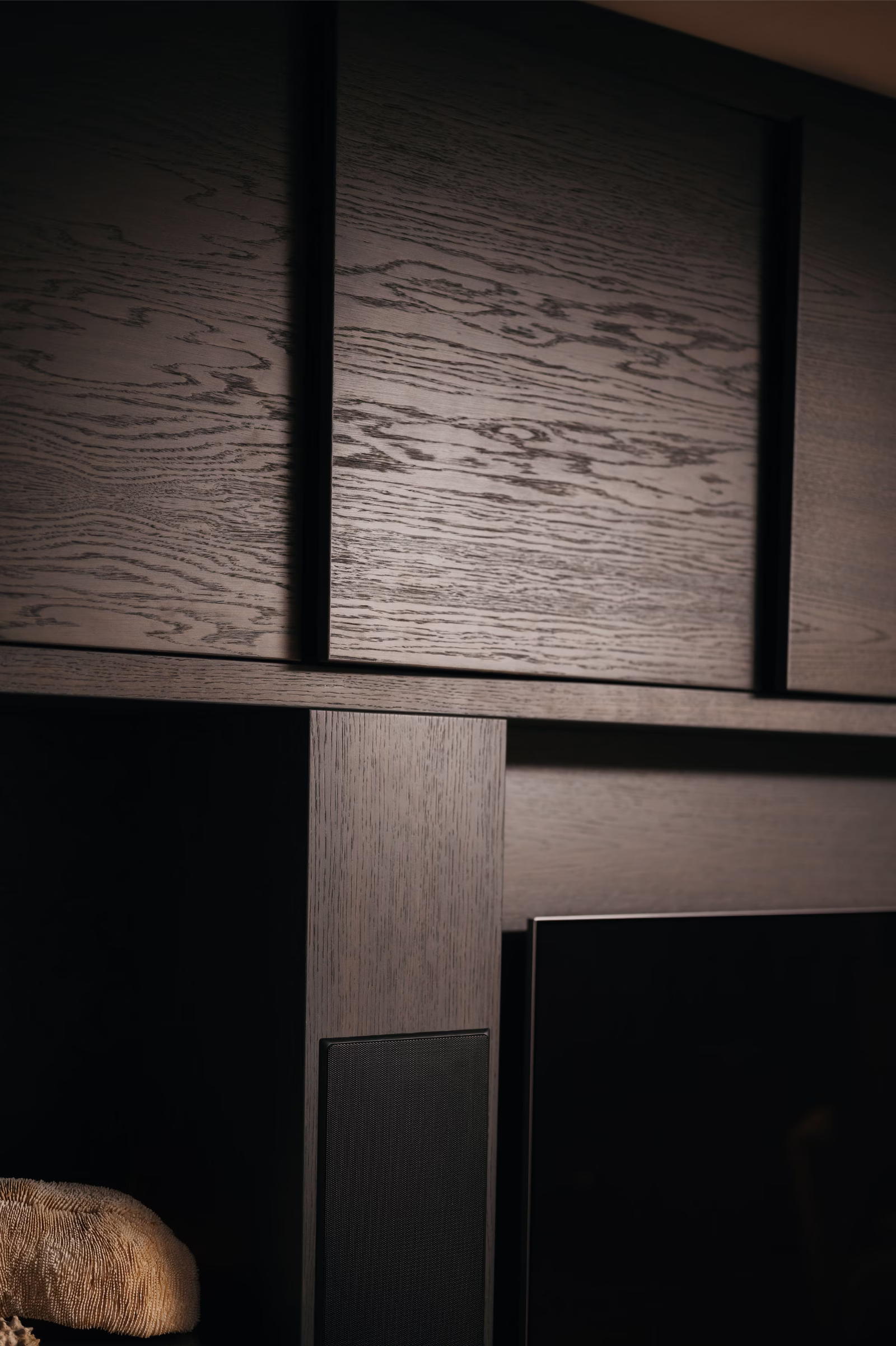 Close-up van donkere houten panelen die een modern, geometrisch wandontwerp vormen met gedefinieerde verticale en horizontale lijnen. De rand van een televisiescherm is zichtbaar in de rechterbenedenhoek.