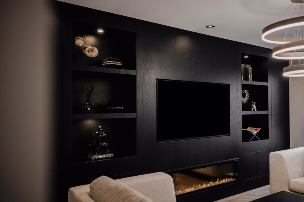 Moderne woonkamer met een strakke zwarte ingebouwde entertainmentwand, planken met decor, een grote tv, een elektrische open haard eronder en beige banken op de voorgrond onder ronde hanglampen.
