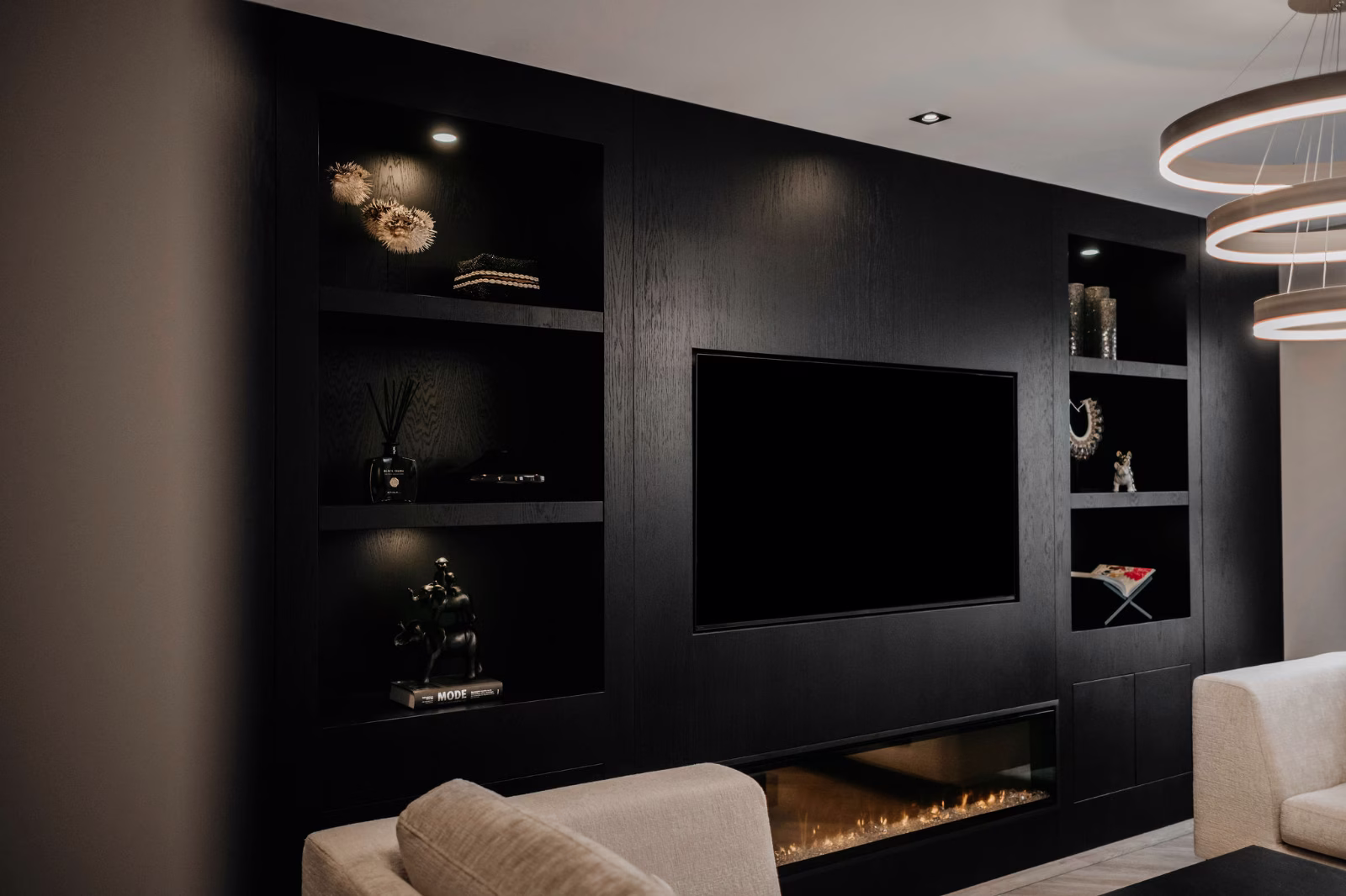 Moderne woonkamer met een strakke zwarte ingebouwde entertainmentwand, planken met decor, een grote tv, een elektrische open haard eronder en beige banken op de voorgrond onder ronde hanglampen.