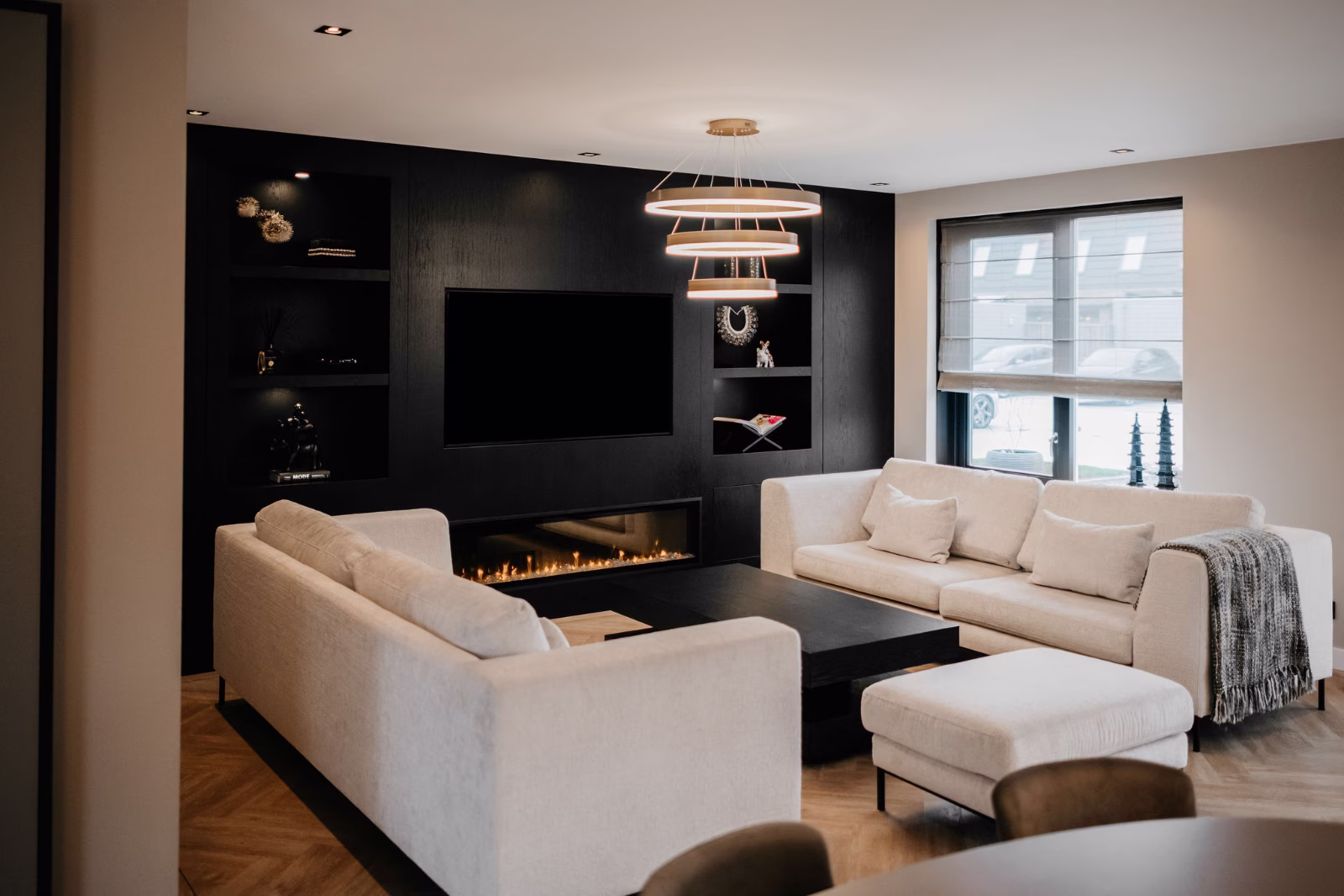 Moderne woonkamer met twee witte banken, een bijpassende voetenbank, een zwarte salontafel, een ingebouwde open haard en een zwarte accentmuur met planken en een tv; groot raam met luxaflex aan de rechterkant.