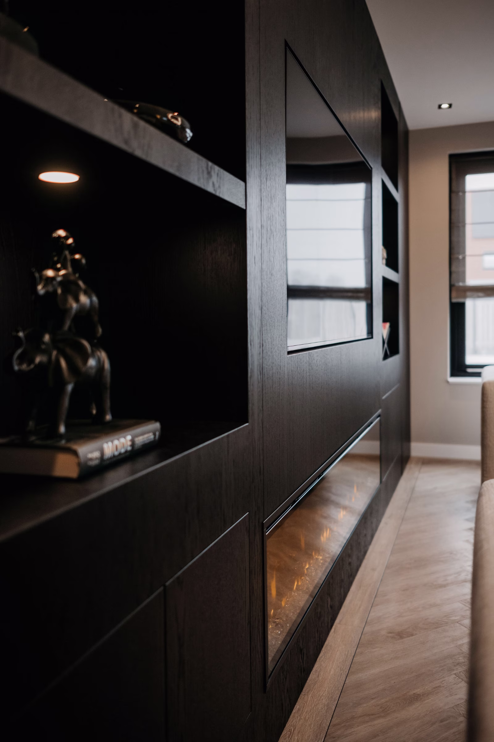 Een moderne woonkamer met donkerhouten ingebouwde rekken, een grote flatscreentelevisie, een elektrische open haard en een beeld op een boek. Lichthouten vloeren en een raam met zwarte jaloezieën zijn zichtbaar.