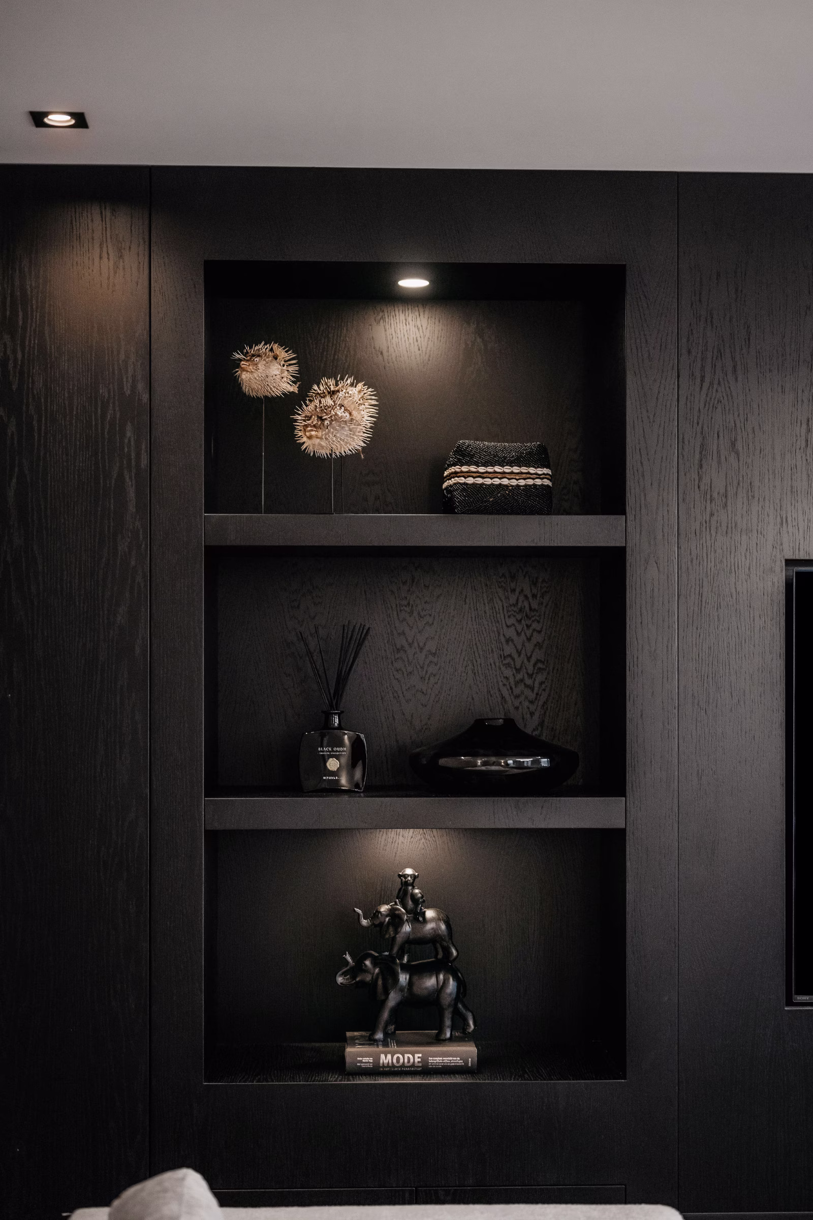 Een modern zwart inbouwrek met drie planken, met decoratieve voorwerpen: twee stekelige bollen en gevouwen textiel op de bovenste plank, een vaas en schaal in het midden en een paardenfiguurtje op boeken op de onderste plank.