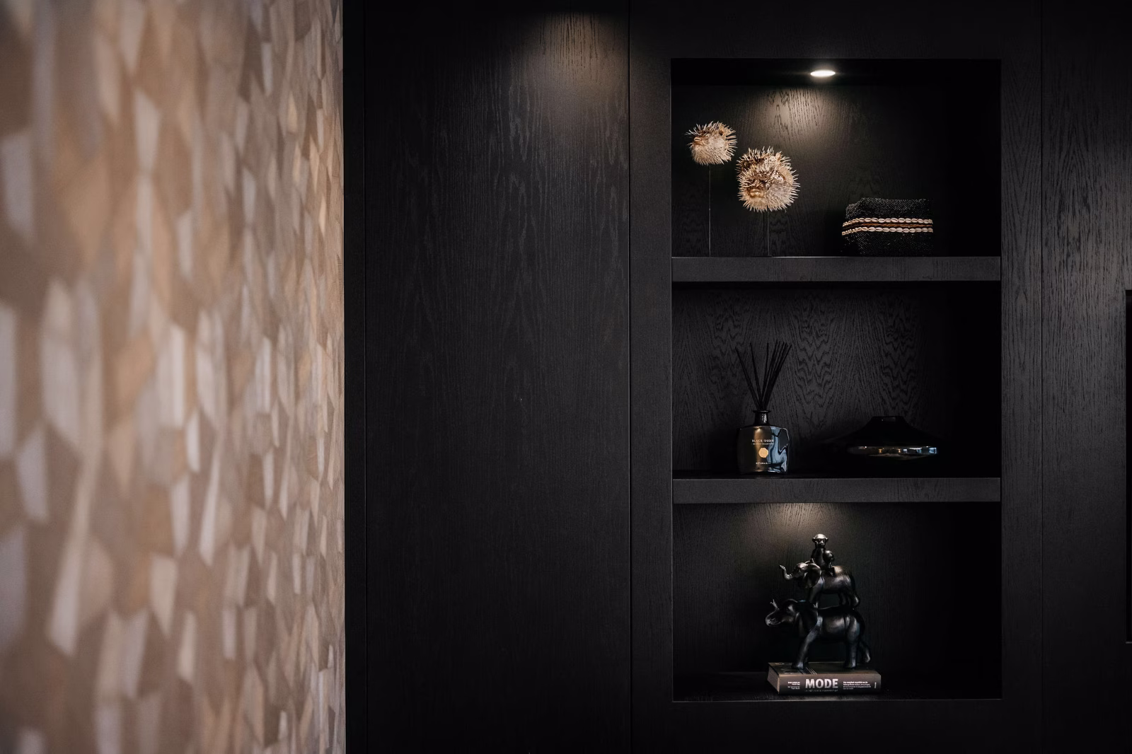 Een moderne, donkerhouten stellingkast met drie planken waarop decoratieve voorwerpen staan, waaronder sculpturale objecten, boeken en geurstokjes; links is een beige muur met geometrisch patroon zichtbaar.