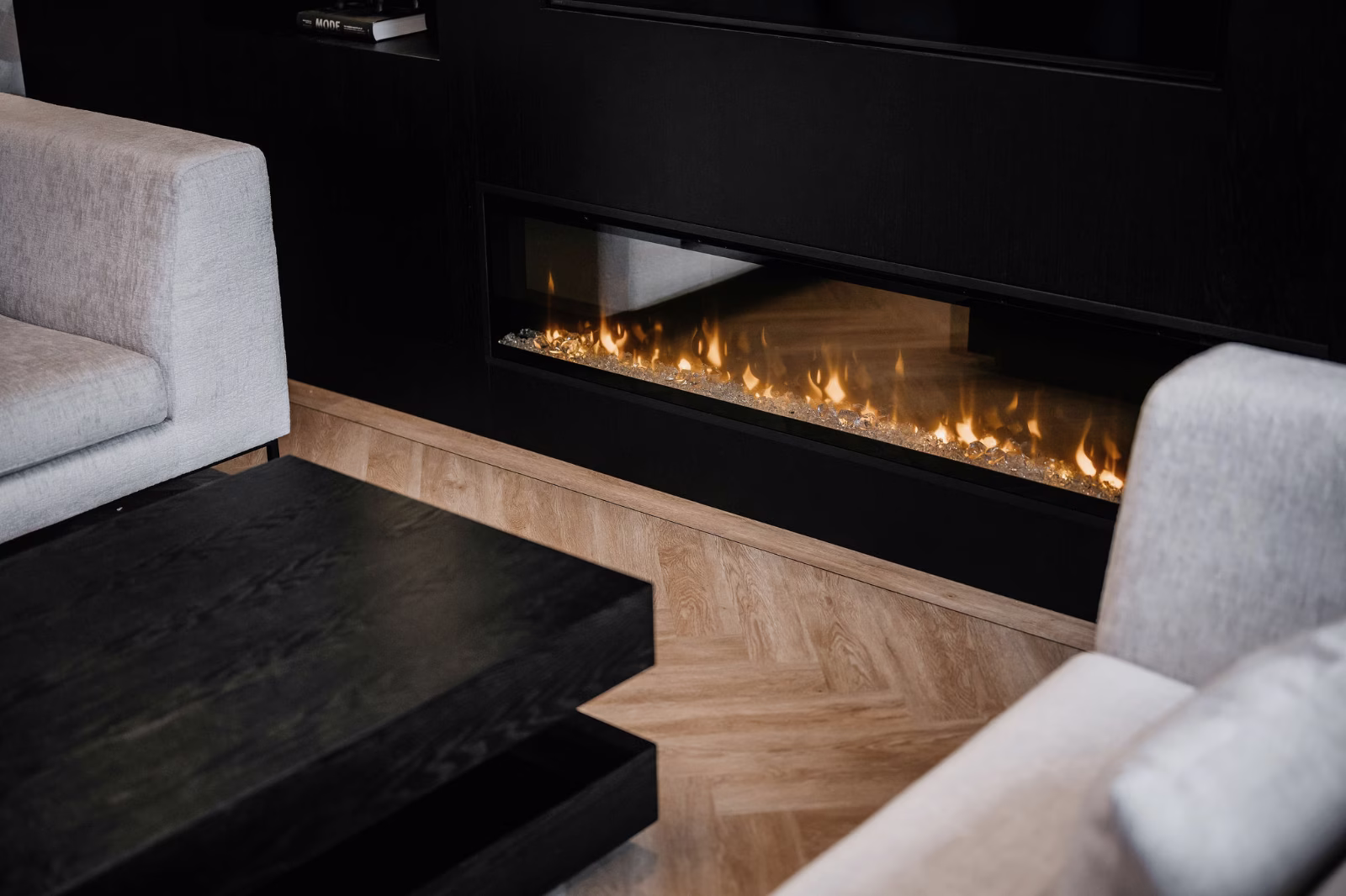 Een moderne woonkamer met lichtgrijze banken, een zwarte salontafel en een strakke inbouwhaard langs een zwarte muur, met vlammen zichtbaar achter glas en een lichthouten vloer.