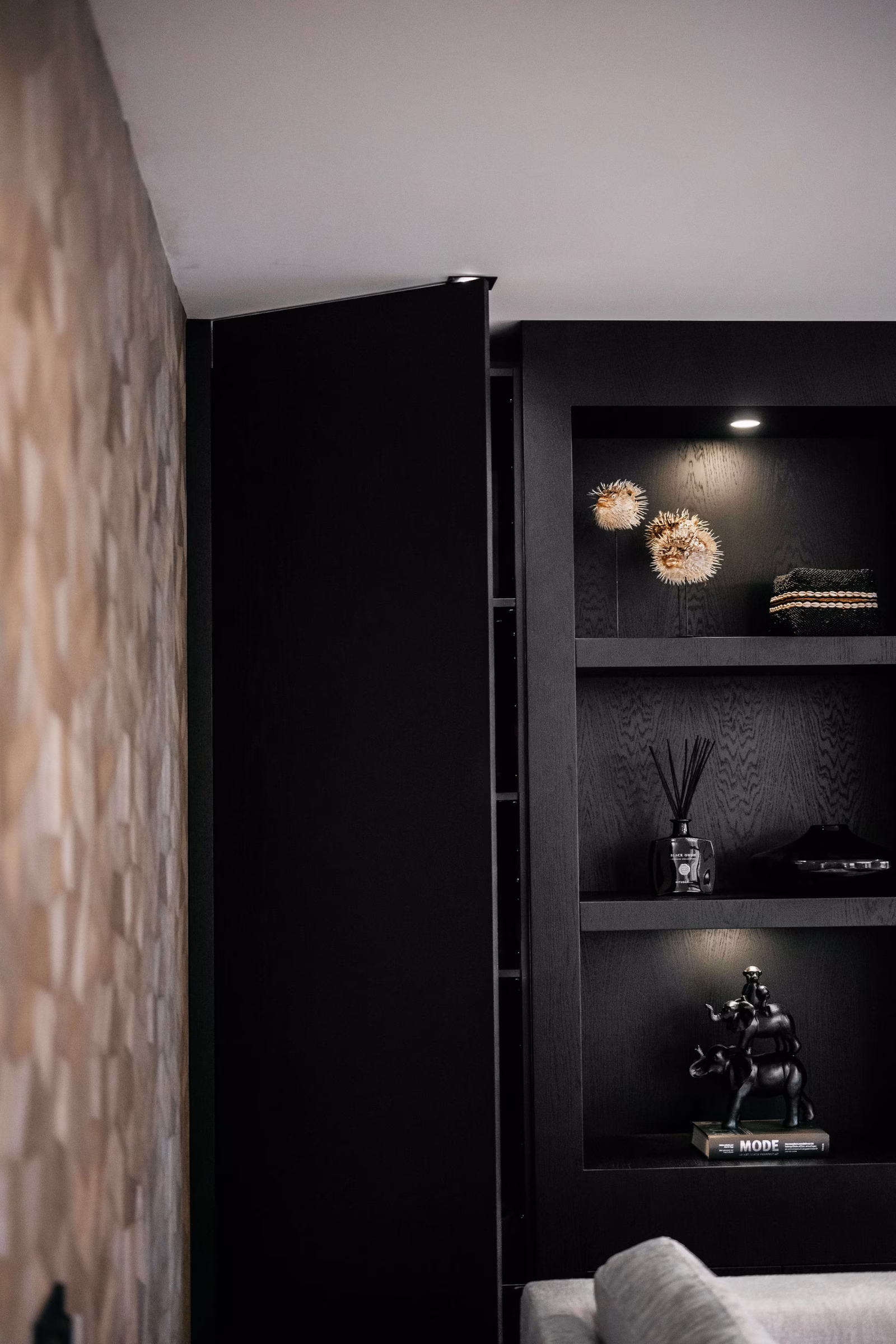 Een moderne zwarte boekenplank met spots toont decoratieve voorwerpen en boeken. Links staat een gedeeltelijk open deur en rechtsonder is een deel van een lichtgrijze bank zichtbaar. De muur links heeft een structuurpatroon.