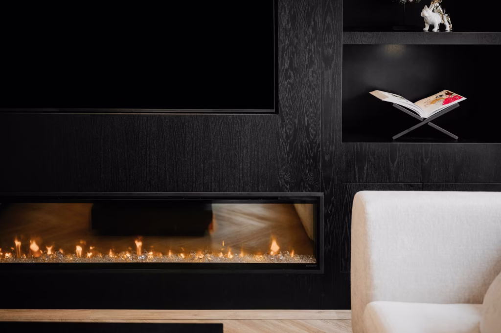 Een moderne leefruimte met een ingebouwde lineaire open haard, zwarte rekken met een decoratieve witte olifant en een open boek, en een deel van een wit gestoffeerde fauteuil op de voorgrond.