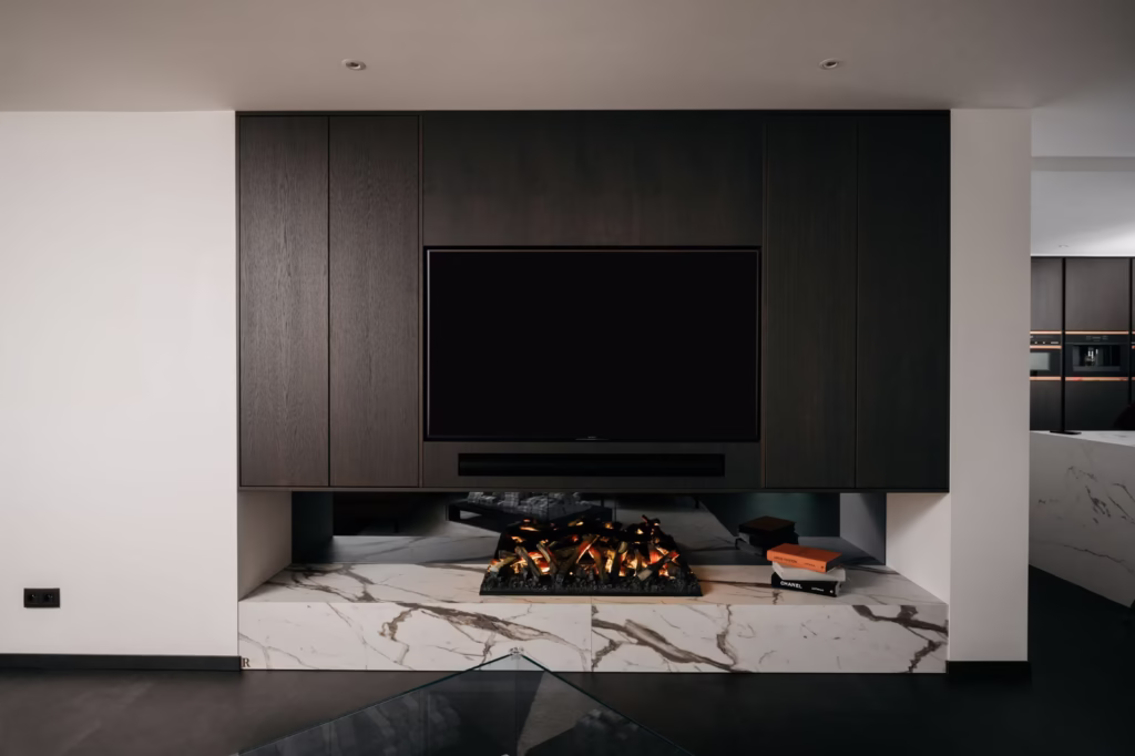 Een moderne woonkamer met een tv aan de muur boven een strakke elektrische open haard, in een donkerhouten lambrisering en wit marmer met grijze aderen; boeken en decorstukken rusten op de marmeren richel.