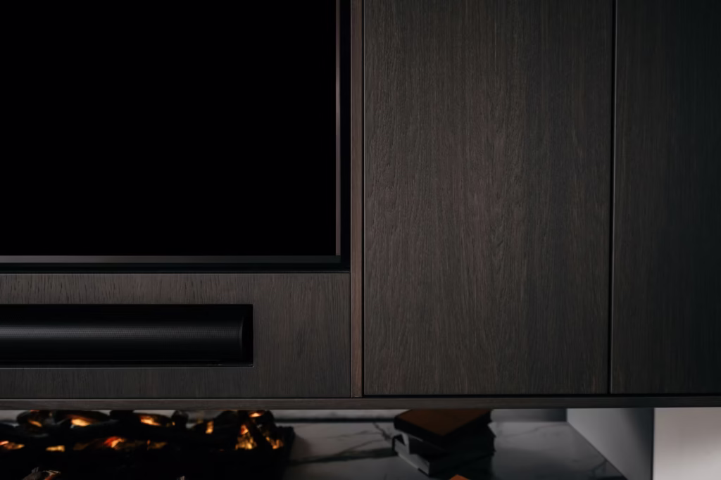Een close-up van een modern donkerhouten entertainmentmeubel met een flatscreentelevisie, een soundbar eronder en een deel van een kunstmatige open haard met gloeiende sintels linksonder. Twee gestapelde boeken zijn gedeeltelijk zichtbaar aan de rechterkant.