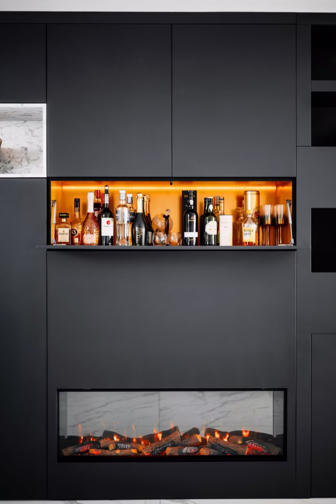 Een modern zwart wandmeubel heeft een ingebouwde elektrische haard met glazen voorkant onder een verlichte plank met flessen drank, cocktailshakers en glaswerk.