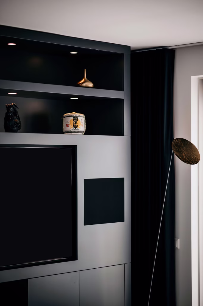 Een moderne, minimalistische woonkamer met donkere ingebouwde planken met decoratieve voorwerpen, waaronder een klein sakevat, een beeld en een gouden tol, naast een flatscreentelevisie en een hoge, dunne staande lamp.