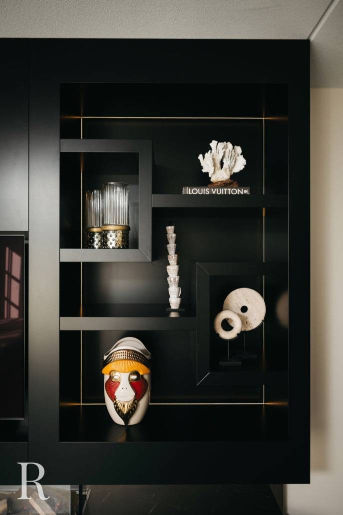 Een modern zwart rek toont verschillende decoratieve voorwerpen, waaronder gestapelde heldere glazen, een wit koraalornament, een Louis Vuitton boek, twee stenen schijven, een kaars en een kleurrijk masker geïnspireerd op tribals.