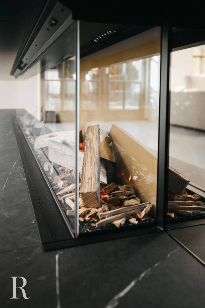 Een moderne, met glas omsloten open haard met gestapeld brandhout en sintels, in een zwart marmeren of stenen ombouw, met een lichte, luchtige kamer zichtbaar op de achtergrond.