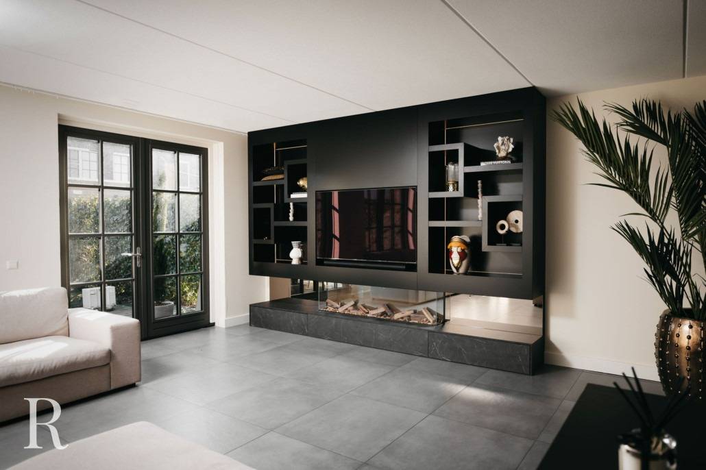 Moderne woonkamer met grote zwarte ingebouwde planken rond een flatscreentelevisie, decoratieve voorwerpen op planken, een beige bank, grote ramen met zwarte kozijnen, grijze tegelvloer en een hoge potplant.