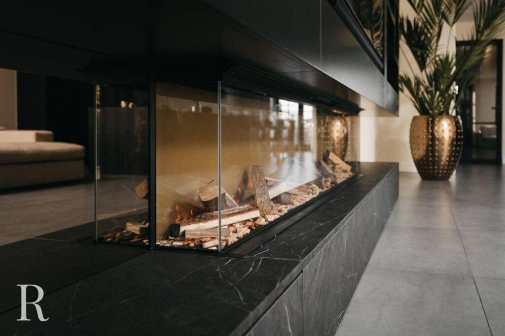 Een moderne binnenhaard met glazen zijkanten heeft brandende houtblokken op een zwart marmeren voet. De kamer heeft strakke, grijze tegelvloeren, een beige bank, een hoge gouden plantenbak en grote groene planten.