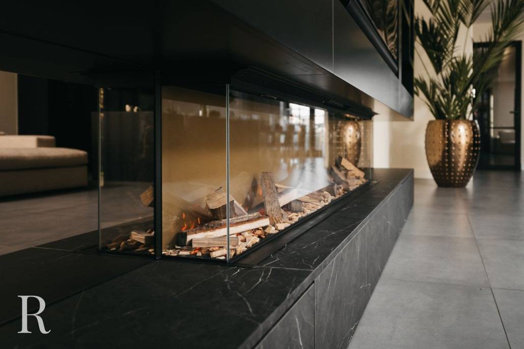 Een moderne binnenhaard met glazen zijkanten heeft brandende houtblokken op een zwart marmeren voet. De kamer heeft strakke, grijze tegelvloeren, een beige bank, een hoge gouden plantenbak en grote groene planten.