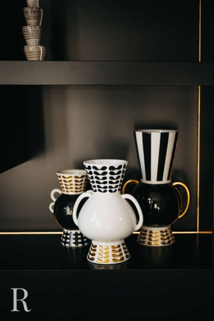 Een groep decoratieve vazen van keramiek met zwarte, witte en gouden geometrische patronen staat op een zwarte plank met gouden rand. De grootste vaas heeft twee gouden handvatten en één vaas heeft verticale zwarte en witte strepen.