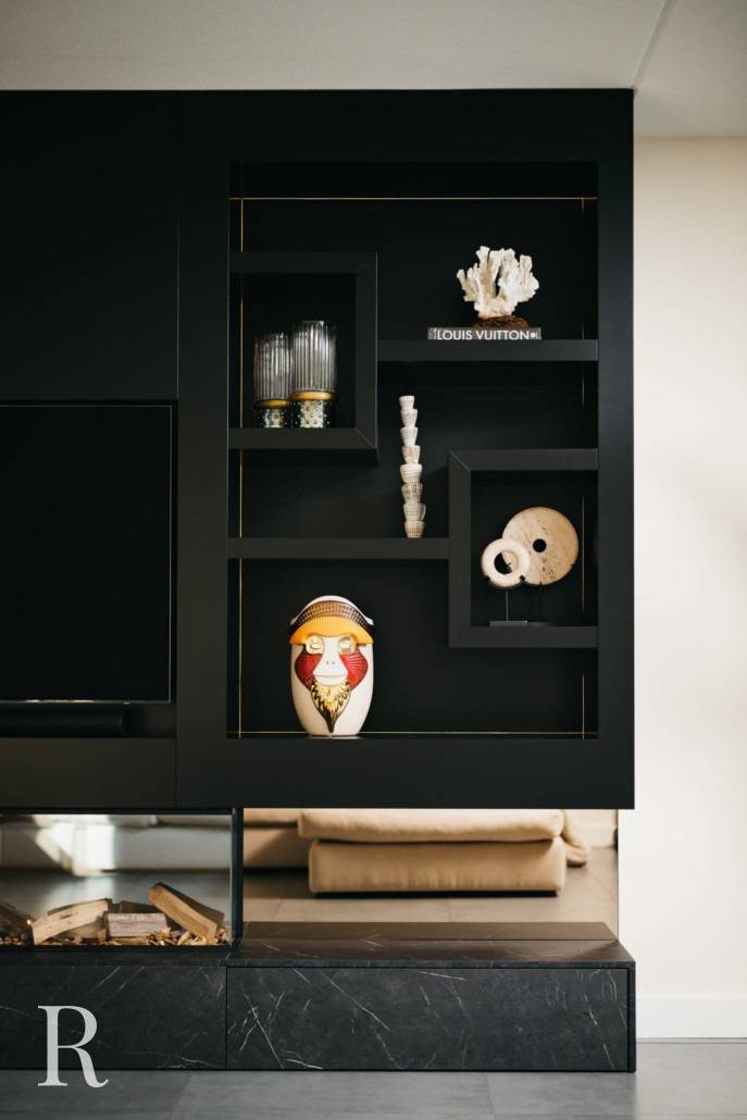 Een modern zwart rek toont verschillende decoratieve voorwerpen, waaronder een vaas, kandelaars, koraal, boeken en maskers, met links een deel van een tv en een open haard.