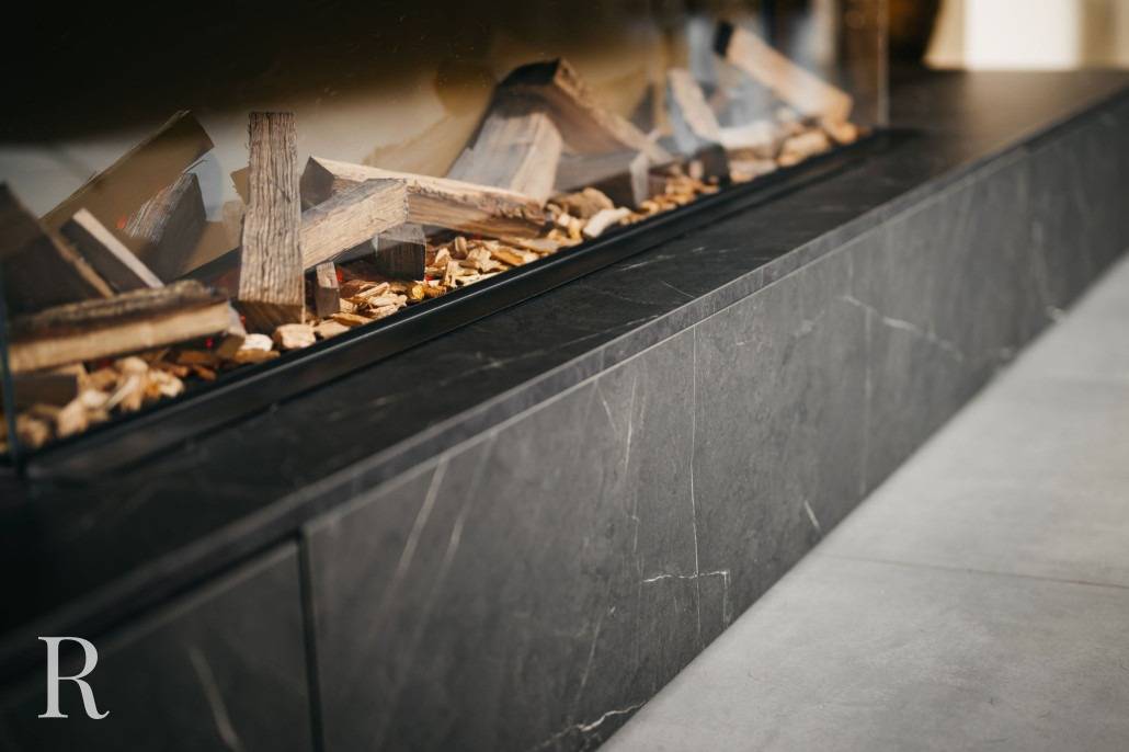 Een close-up van een moderne open haard met gestapelde houtblokken en houtsnippers binnenin, in een strakke, donkere marmeren of stenen ombouw. Het ontwerp is minimalistisch en eigentijds.