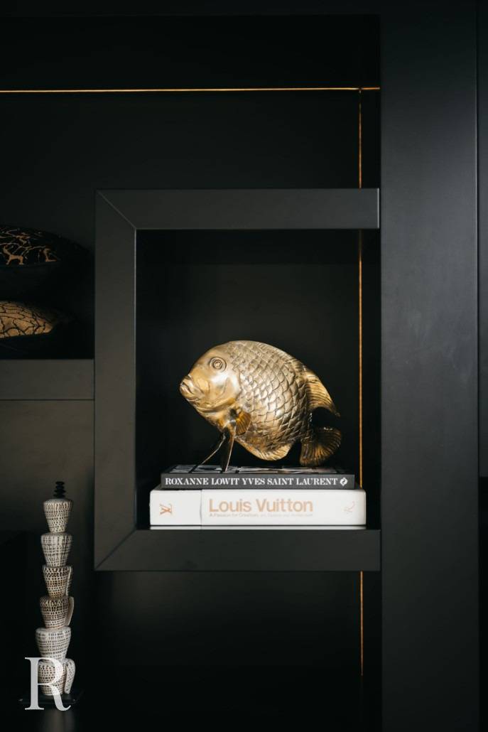 Een metallic gouden vissculptuur staat op een plank, bovenop twee gestapelde boeken, waarvan één met het label Louis Vuitton. De achtergrond is een matzwarte kast met minimalistisch decor.