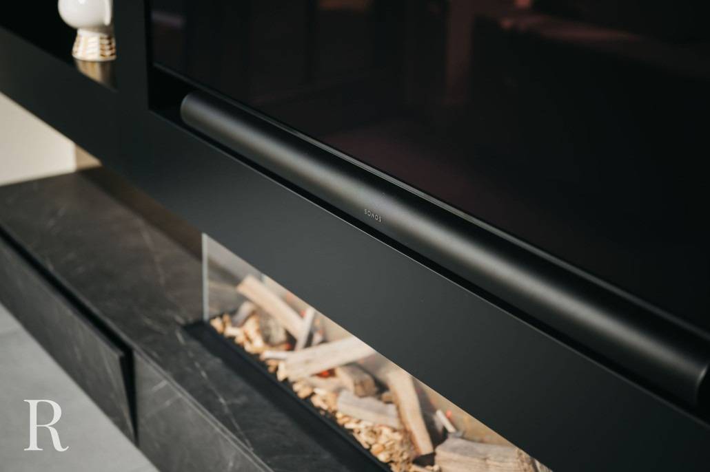 Een close-up van een moderne zwarte soundbar onder een flatscreen-tv, met een ingebouwde elektrische open haard en decoratieve houtsnippers onder het tv-meubel.