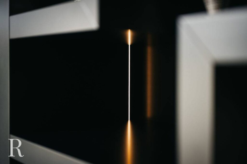 Een close-up van geometrische planken met een smalle verticale opening. Warm licht schijnt door de opening en werpt een gloeiende lijn en reflectie op de donkere binnenkant. De hoek van een witte "R" is linksonder zichtbaar.