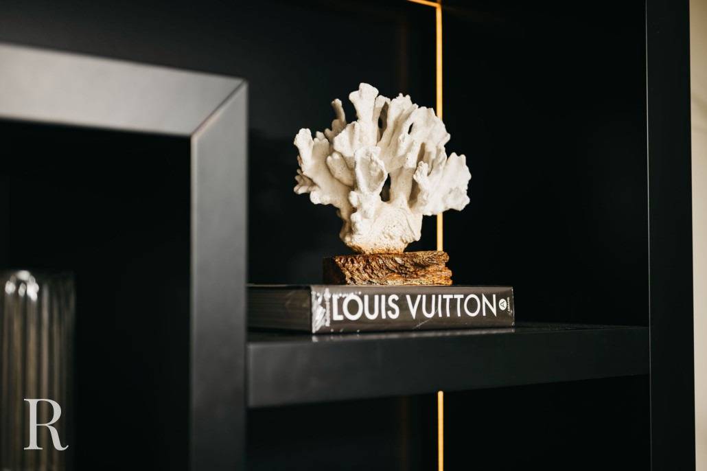 Een wit koraalornament ligt bovenop een Louis Vuitton koffietafelboek, uitgestald op een modern zwart rek met een zacht licht dat de items benadrukt.