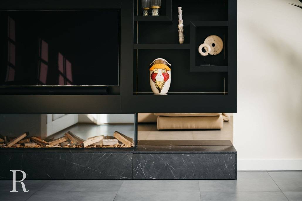 Een modern zwart rek toont decoratieve voorwerpen, waaronder een kleurrijk uilenbeeldje en een sculptuur met textuur. Hieronder ligt gestapeld brandhout achter een glazen paneel naast een zwarte marmeren voet.