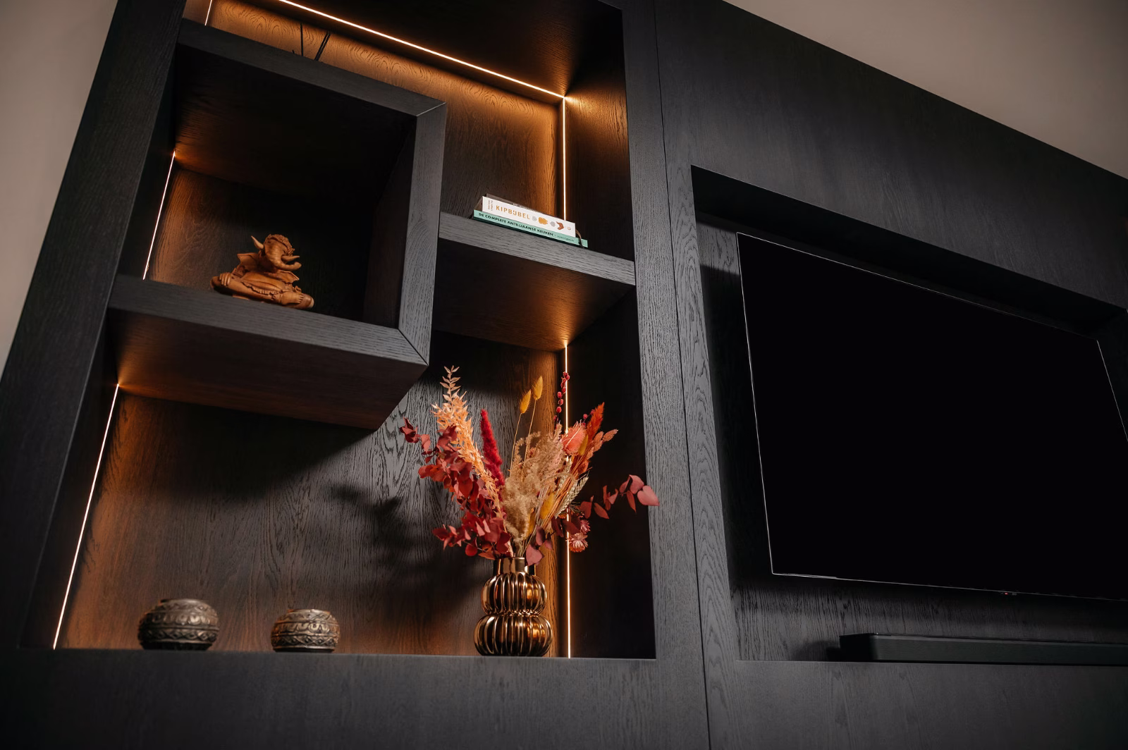 Een modern donkerhouten wandmeubel met ingebouwde planken bevat een tv, een klein Boeddhabeeld, een paar boeken, decoratieve vazen en een boeket gedroogde bloemen, allemaal zacht verlicht door warme accentverlichting.