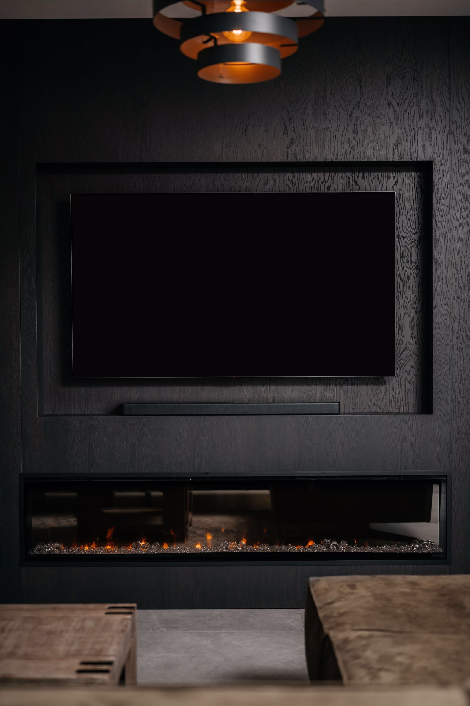 Een moderne woonkamer heeft een flatscreentelevisie op een donkere muur met textuur boven een strakke, lineaire elektrische open haard. Daarboven hangt een eigentijdse plafondlamp en op de voorgrond is een deel van een beige bank te zien.