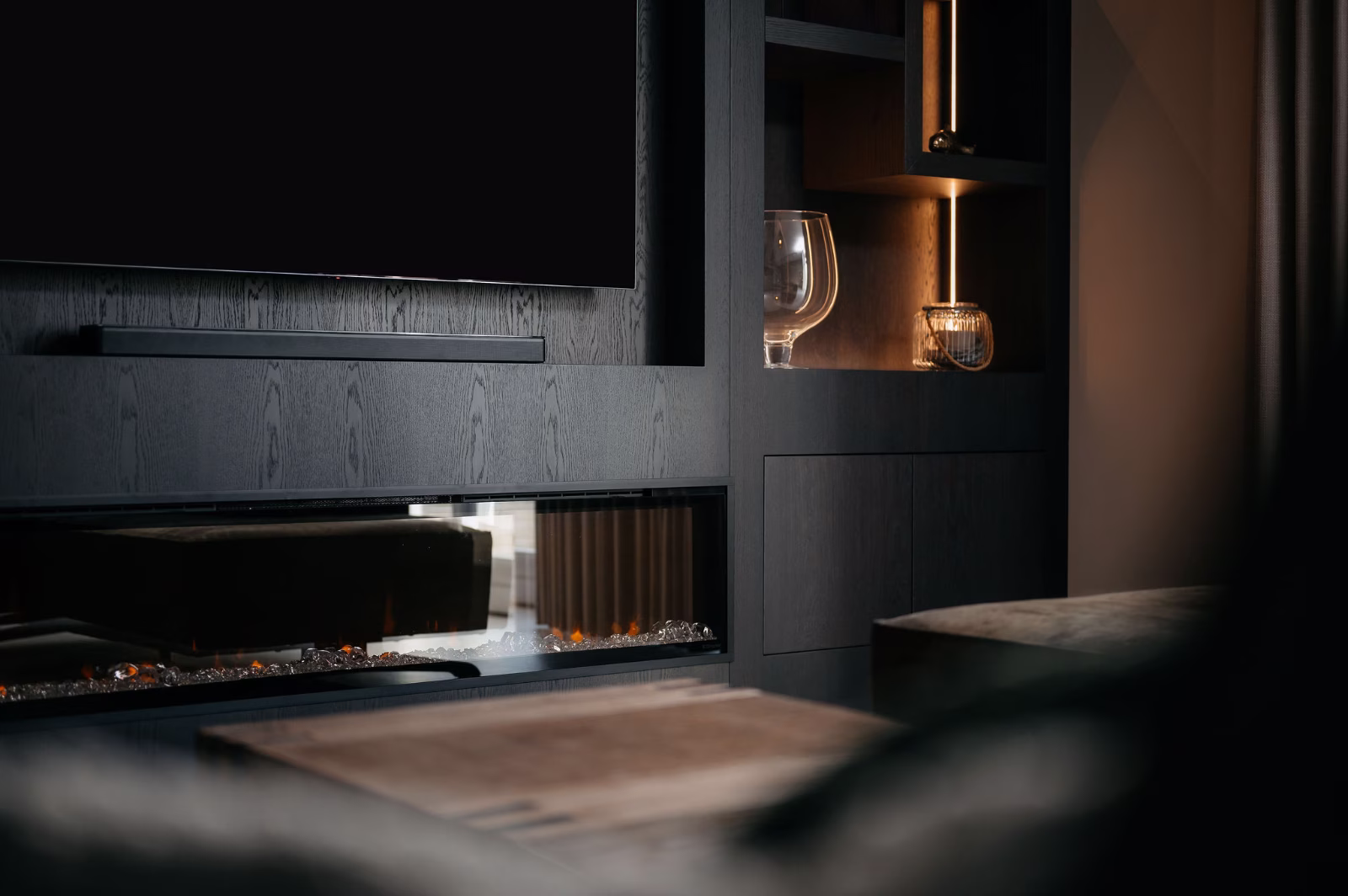 Een moderne woonkamer heeft een strak zwart entertainmentmeubel met een tv aan de muur, een elektrische open haard eronder, glaswerk op planken en zachte, warme verlichting die een gezellige sfeer creëert.