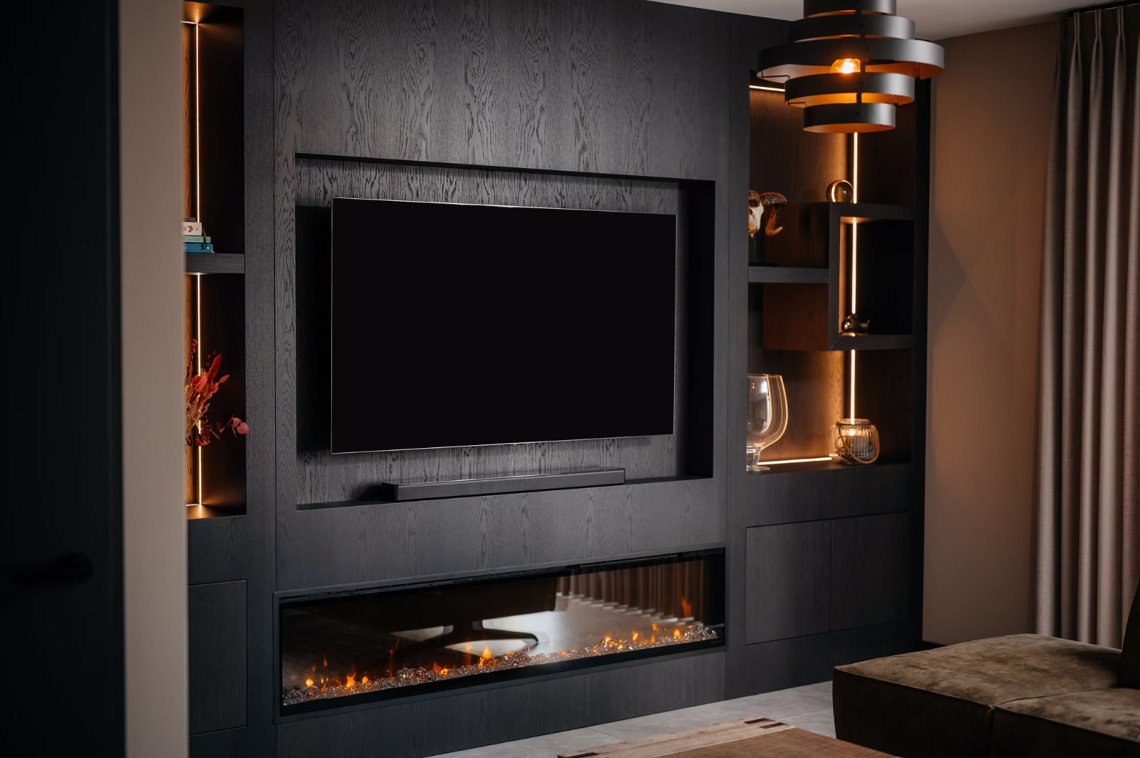 Een moderne woonkamer heeft een strakke zwarte entertainmentwand met een gemonteerde tv, ingebouwde planken met decoratieve voorwerpen en een elektrische open haard onder de tv. Warme verlichting voegt een gezellige sfeer toe.