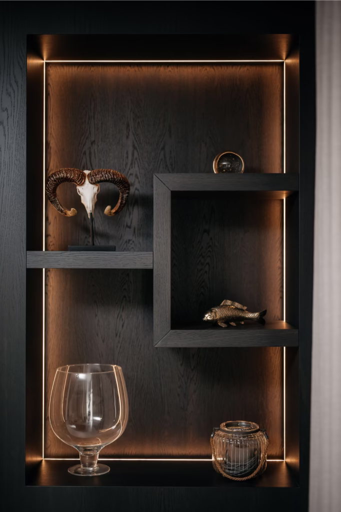 Op een donkere houten plank met ingebouwde warme verlichting staan een ramsschedel, een klein gouden visbeeldje, een glazen bol, een grote lege glazen vaas en een geribbelde glazen pot.