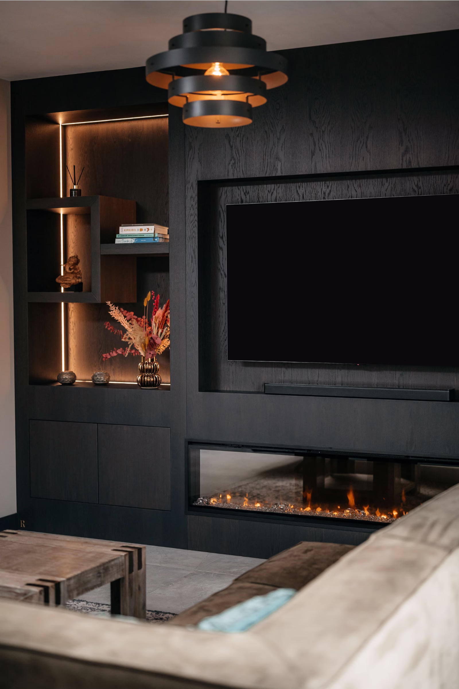 Moderne woonkamer met donkerhouten ingebouwde planken, een flatscreentelevisie, een elektrische open haard eronder, decoratieve voorwerpen, boeken, een vaas met bloemen, warme verlichting en een hedendaagse plafondlamp.