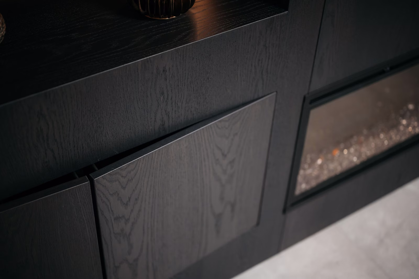 Close-up van een moderne, donkerhouten kast met een gedeeltelijk open deur en een compartiment met glazen voorkant rechts, tegen een lichtgekleurde vloer.