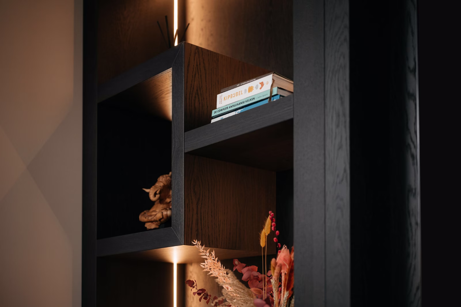 Een moderne donkerhouten plank met ingebouwde verlichting, met een paar gestapelde boeken, een decoratief houten voorwerp en een bos gedroogde bloemen in het onderste vak.