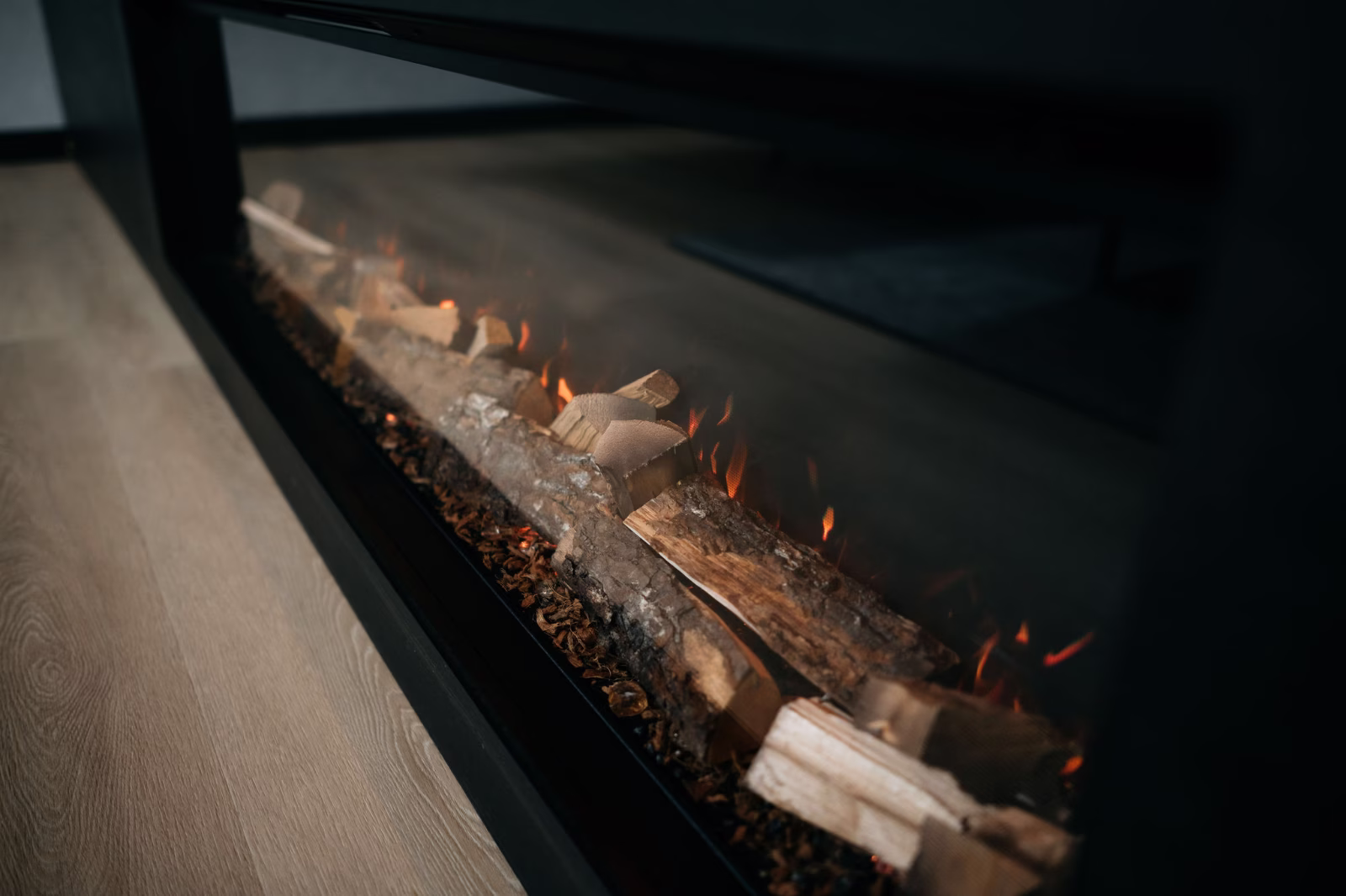 Een moderne elektrische open haard met realistisch ogende houtblokken en gloeiende oranje vlammen, in een strak zwart frame, is ingebouwd in een muur boven een lichte houten vloer.
