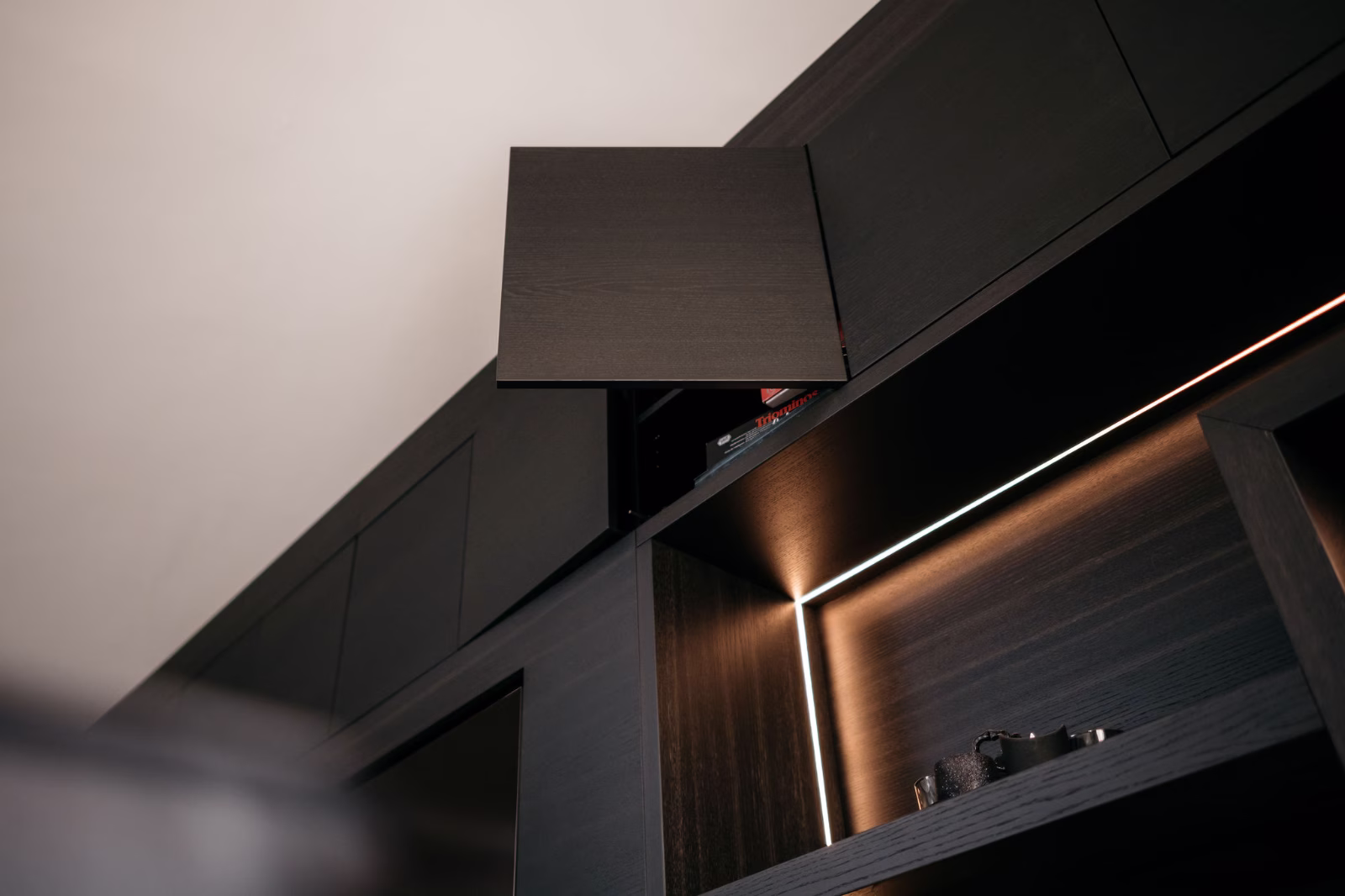 Een donkere houten kast met een open bovendeur die een gedeeltelijk verborgen plank onthult. Warme LED-verlichting benadrukt de onderste open plank, die een paar kleine voorwerpen bevat. De foto is genomen vanuit een lage hoek.