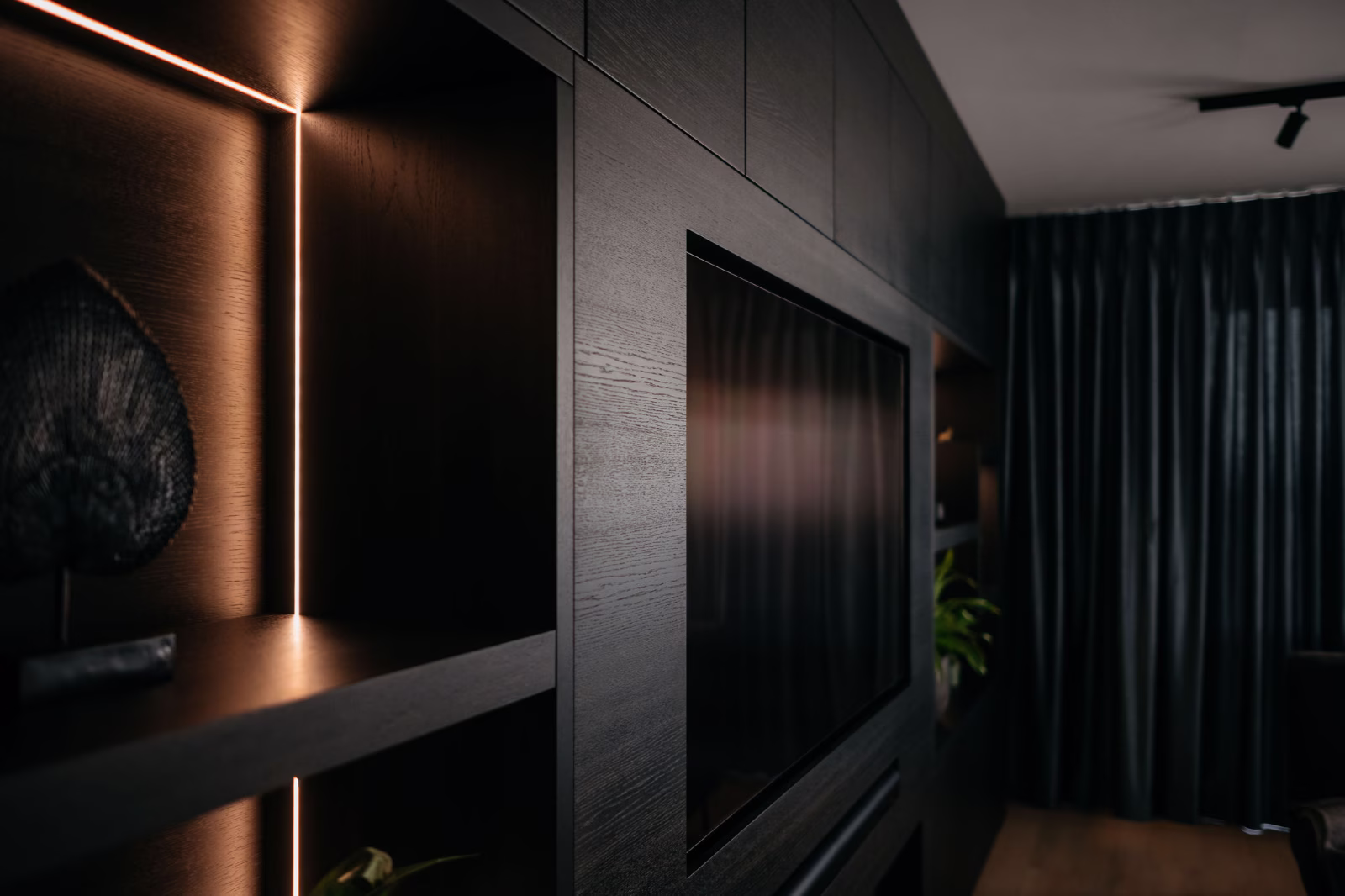 Een moderne woonkamer met een donker houten entertainmentmeubel met ingebouwde planken, subtiele inbouwverlichting, een tv aan de muur en donkere gordijnen van vloer tot plafond.