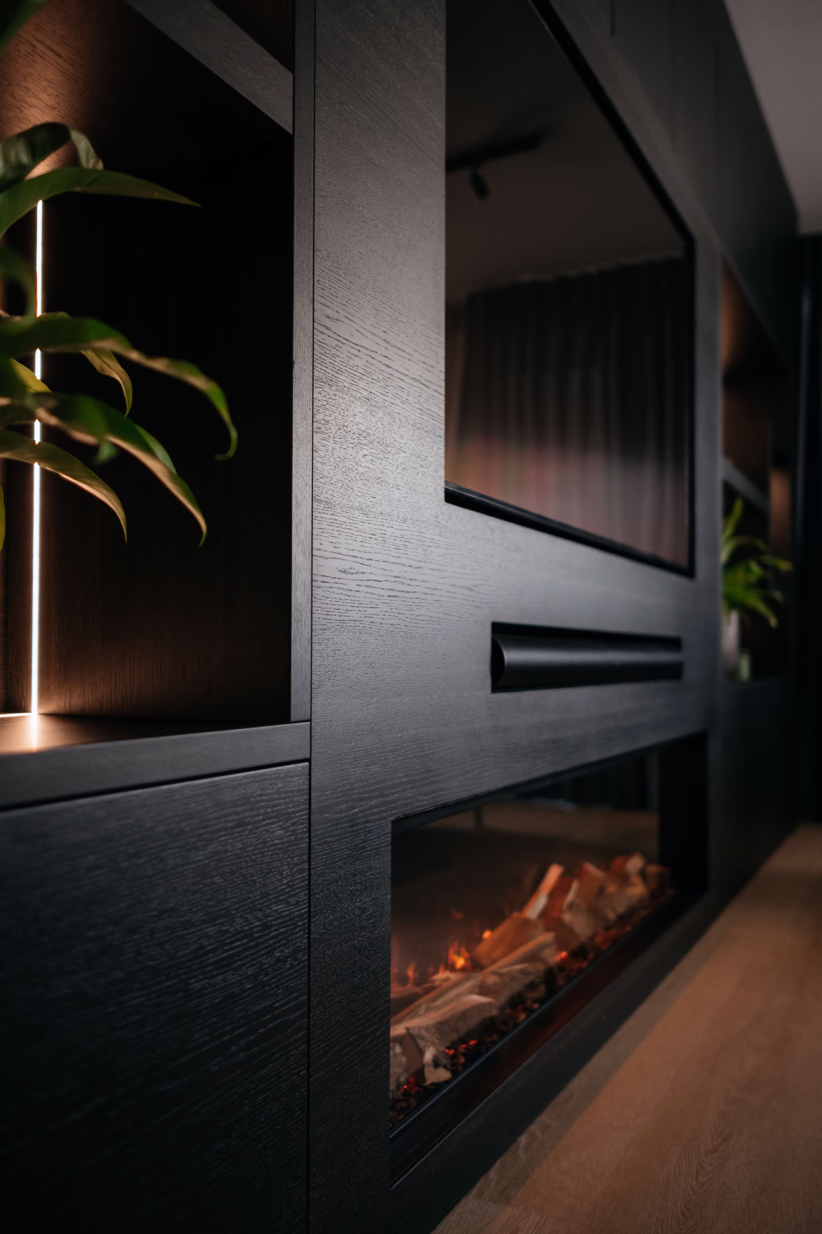 Een modern zwart wandmeubel met ingebouwde planken, een grote tv, kamerplanten en een elektrische open haard met gloeiende sintels in een stijlvolle, schemerig verlichte woonkamer.