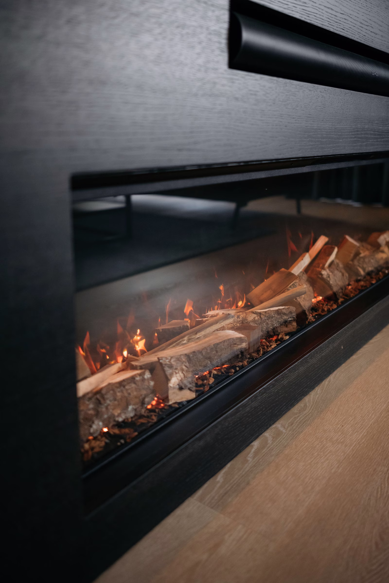 Een close-up van een moderne binnenhaard met brandende houtblokken en gloeiende sintels, omgeven door een strak zwart frame, tegen een houten vloer.