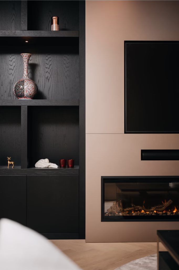 Een moderne woonkamer met ingebouwde zwarte rekken met decoratieve spullen, een beige muur met een flatscreentelevisie en een elektrische open haard eronder.
