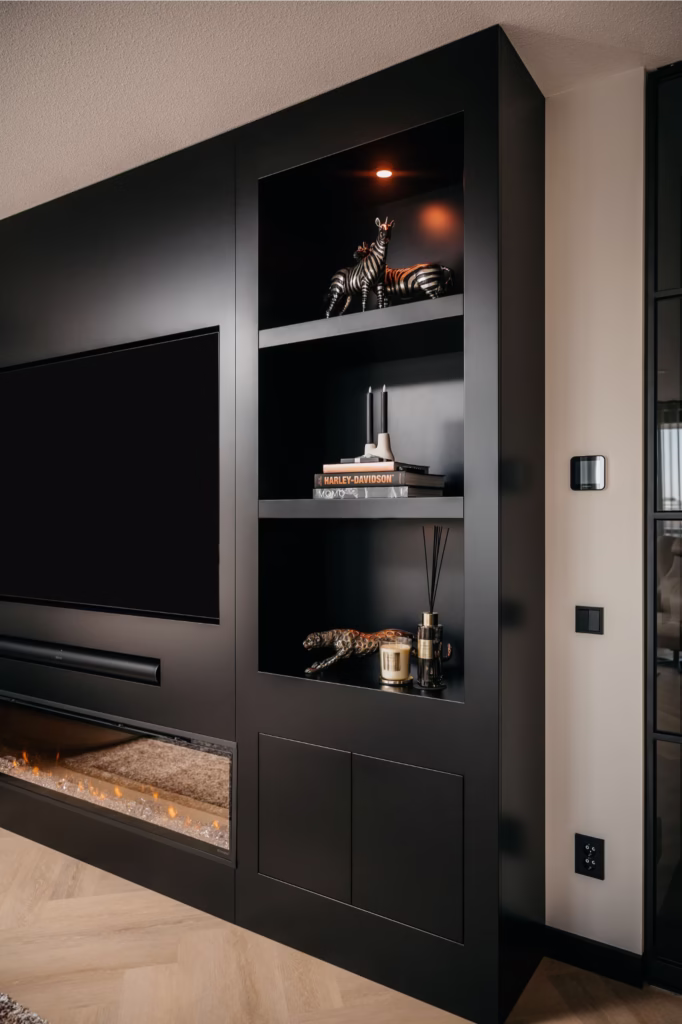 Een moderne, zwarte inbouwkast met decoratieve voorwerpen, boeken en een Harley-Davidson-model, naast een flatscreentelevisie en een elektrische open haard met gloeiende sintels eronder, op een lichthouten vloer.