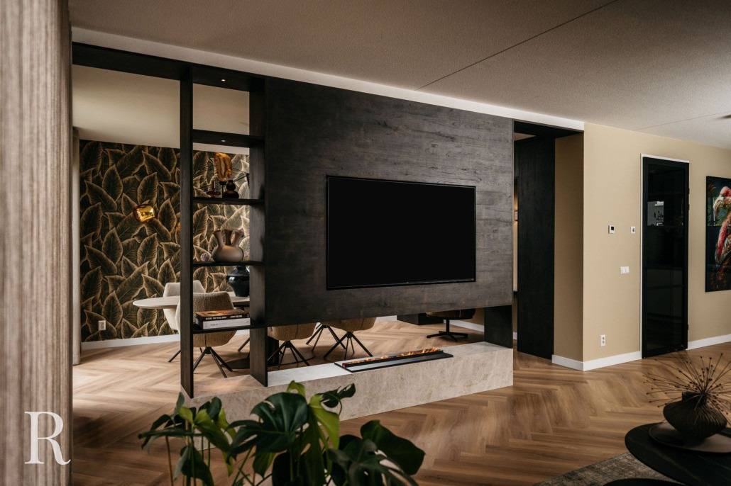 Een moderne woonkamer met een donkerhouten cinewall met een gemonteerde flatscreentelevisie, open rekken, vloeren met houtpatroon en een stijlvolle inrichting, inclusief planten en eigentijdse meubels.