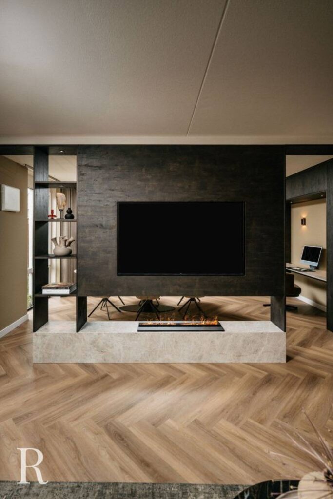Moderne woonkamer met een donkerhouten cinewall met tv aan de muur en ingebouwde lineaire open haard eronder, open planken met decor aan de linkerkant, lichte visgraat houten vloer en een gedeeltelijk zicht op een bureau op de achtergrond.