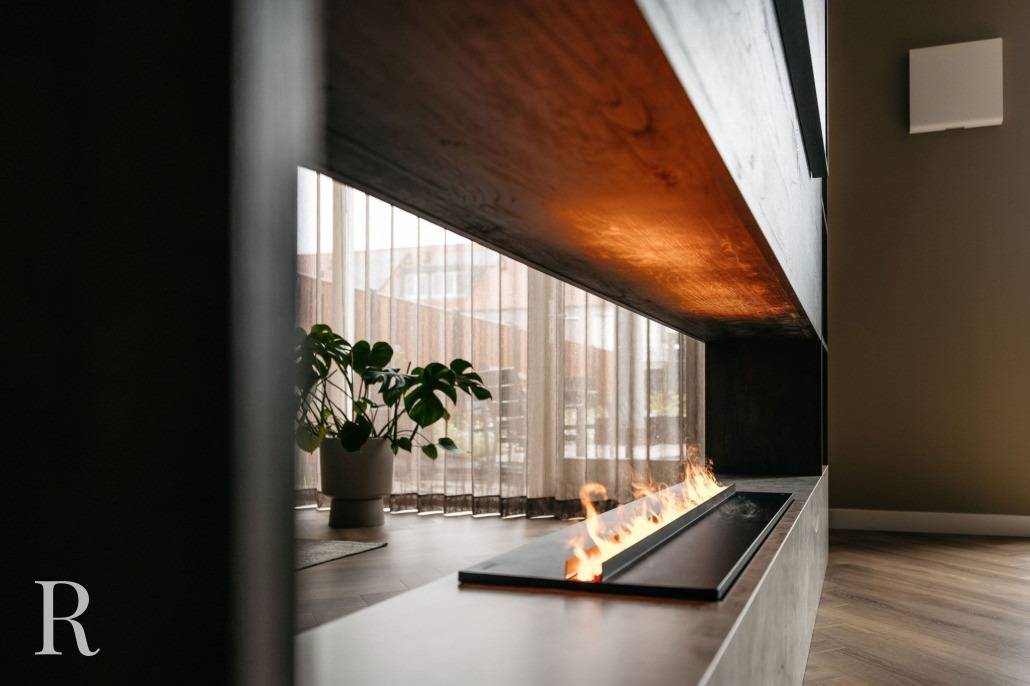 Een moderne lineaire open haard met een zichtbare vlam staat in een strakke, minimalistische woonkamer met een donkerhouten cinewall, grote ramen, transparante gordijnen, een houten vloer en een potplant op de achtergrond.