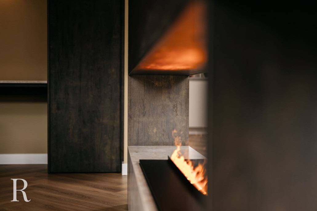 Een close-up van een moderne, ingebouwde open haard in een strakke donkerhouten cinewall met zichtbare vlammen. De kamer heeft een houten vloer en een minimalistisch design. Een gestileerde letter R verschijnt in de linker benedenhoek.