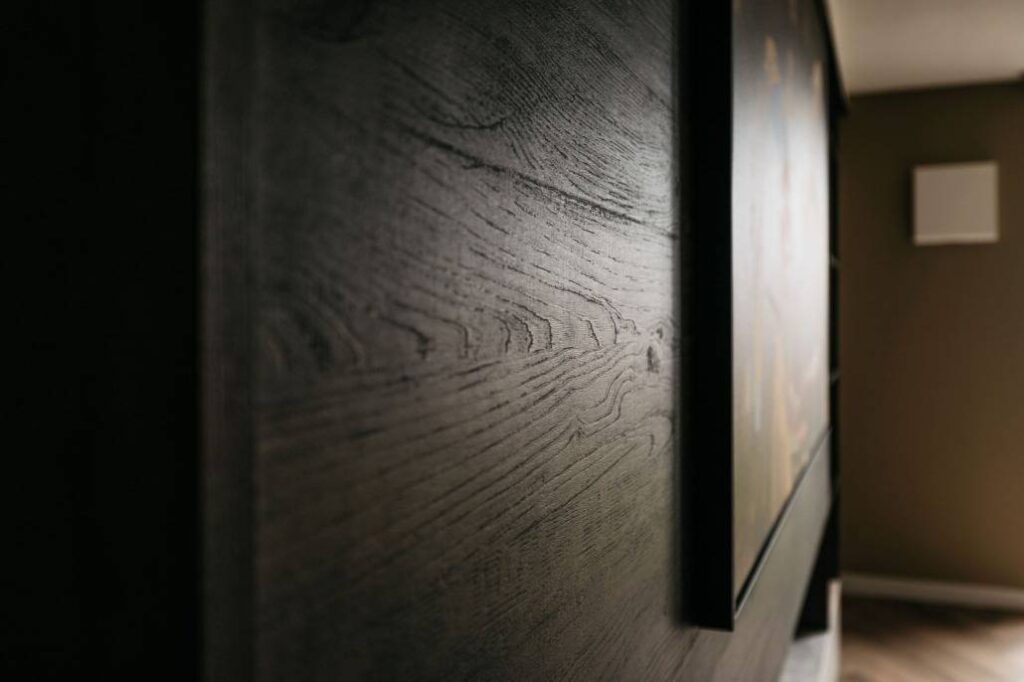 Close-up van een donkerhouten cinewall met zichtbare nerfstructuur, naast een ingelijste foto. De achtergrond is onscherp en toont een deel van een kamer met gedempte verlichting en bruine muren.