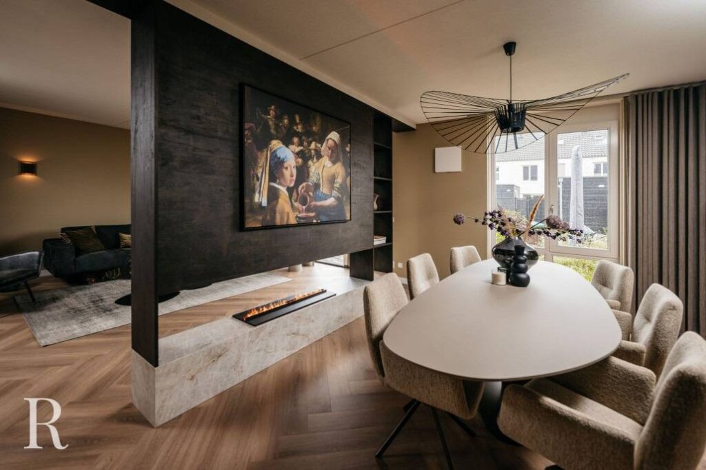 Moderne open woon- en eetruimte met neutrale tinten, ovale eettafel, gestoffeerde stoelen, een Donkerhouten cinewall met elektrische open haard, hedendaagse kroonluchter, groot kunstwerk, houten vloeren en een raam met uitzicht op de tuin.